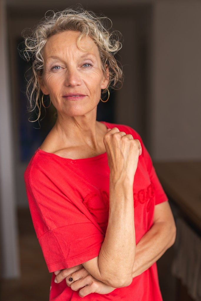 Portrait of Manuela Stoerzer, silver-haired actor, model, and awareness-based coach, wearing a red T-shirt, looking calm and present